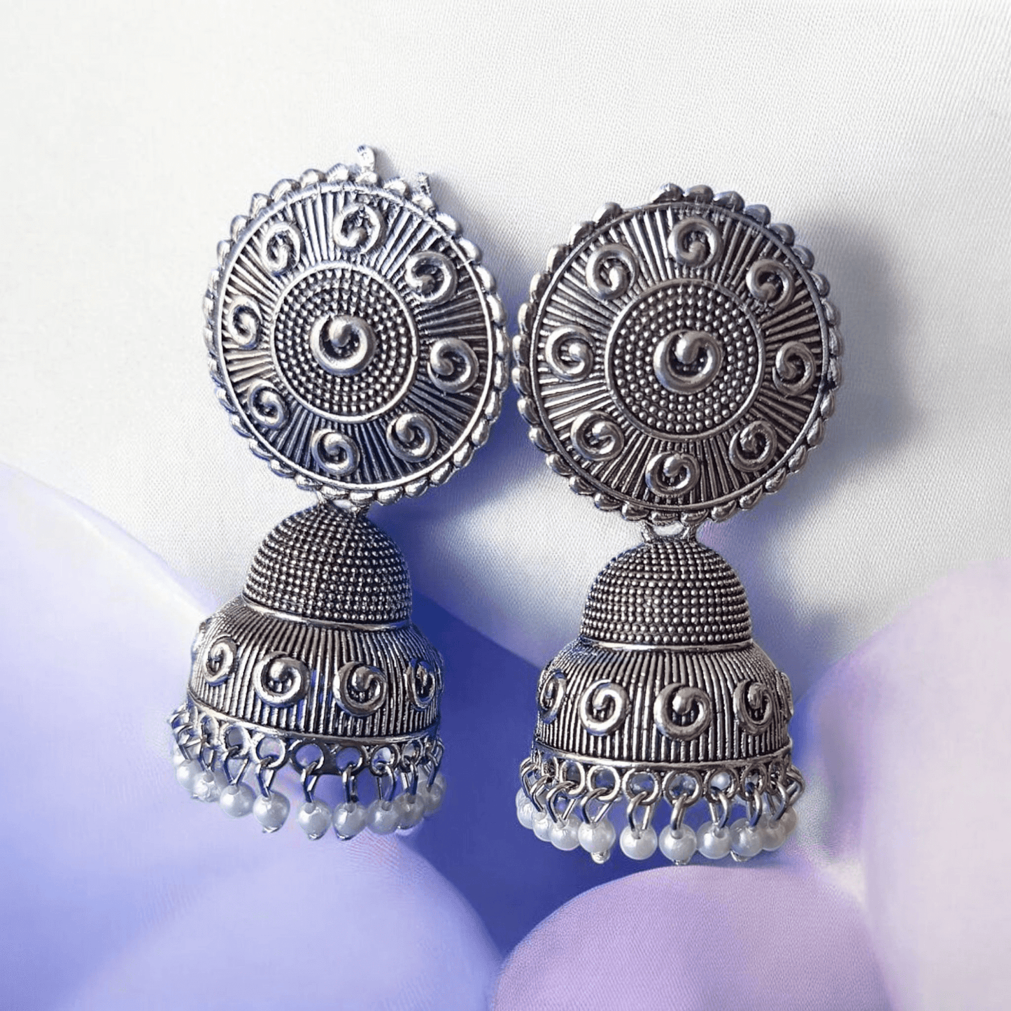 Handcrafted oxidized silver jhumka earrings with intricate designs and pearl drops against a fabric background