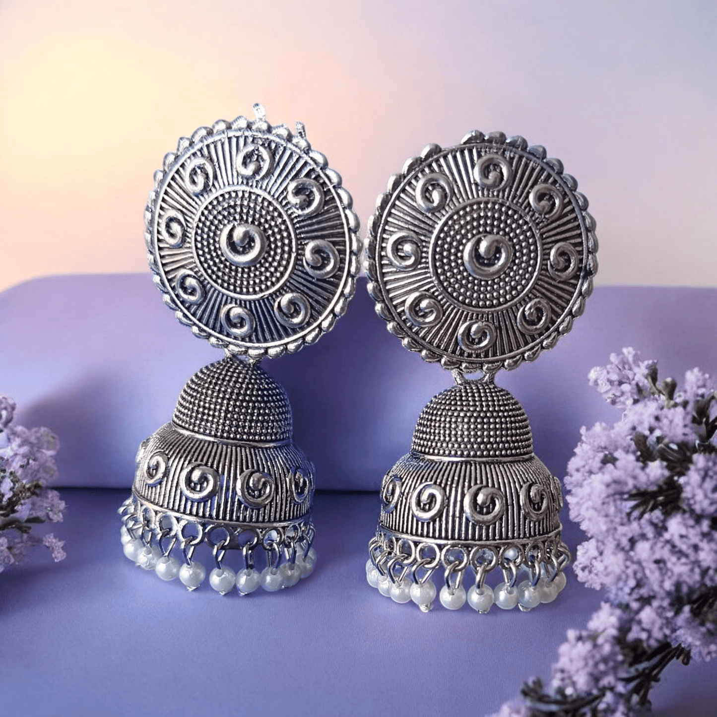 Handcrafted oxidized silver jhumka earrings with intricate designs and pearl drops against a lavender flower and fabric background